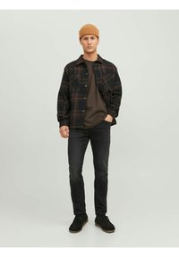 Camicia in flanella nera e a quadri sopra una maglietta scura, abbinata a jeans scuri e scarpe nere. Cappello di lana marrone in testa.