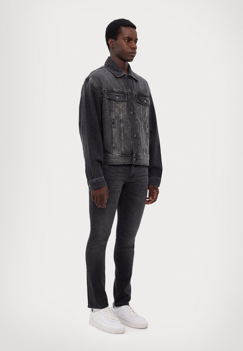 Black denim jacket with button closure, two front pockets, and a slightly faded finish, paired with dark jeans and white sneakers.