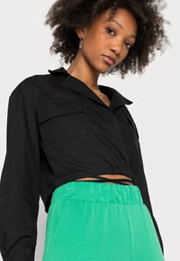 Camisa preta cropped com colarinho e dois bolsos frontais; usada com calças verdes de cintura elástica. Tecido suave e ajuste descontraído.