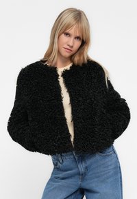 Μαύρο, αφράτο, κοντό μπουφάν με υφή εξωτερικά, στρογγυλή λαιμόκοψη και ανοιχτό μπροστινό μέρος. Φοριέται πάνω από ένα ελαφρύ τοπ και συνδυάζεται με μπλε τζιν.