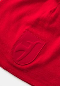 Czerwona dzianinowa czapka beanie z teksturowanym, wypukłym naszywką logo na środku. Materiał jest gładki z ściśle utkanym wzorem i gładkimi brzegami.