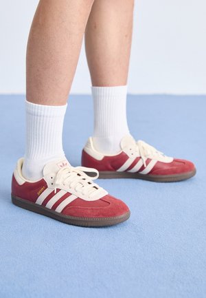 Osoba nosząca czerwone buty sportowe Adidas Samba z białymi paskami i białe skarpetki, stojąca na jasnoniebieskim dywanie.