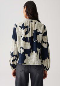 Vrouw met lang donker haar, gekleed in een losse, lange mouwen bloes met bloemenpatroon in marineblauw en beige, gecombineerd met donkergrijze jeans, met haar rug naar de camera gekeerd.