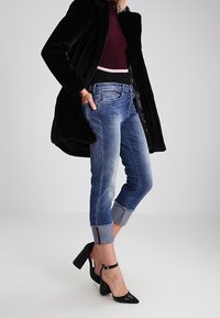 Nainen pukeutuneena mustaan turkkiin, viininpunaisiin neuleisiin, käärittyihin sinisiin farkkuihin ja mustiin nilkkaremmikenkiin, seisoo yksinkertaista taustaa vasten.