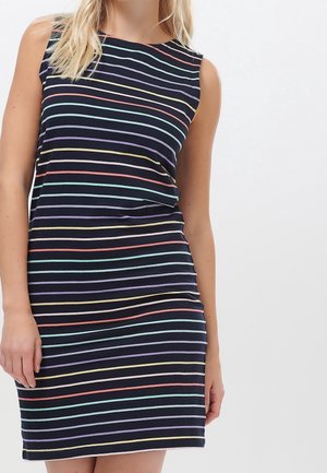 Kobieta ubrana w granatową, bezrękawnikową sukienkę z poziomymi pastelowymi paskami w kolorach różowym, żółtym, fioletowym i jasnoniebieskim na białym tle.