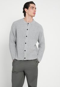 Grå, ribbestrikket cardigan med rund hals, med knappelukking foran og fem mørke knapper. Brukt over olivengrønne bukser.