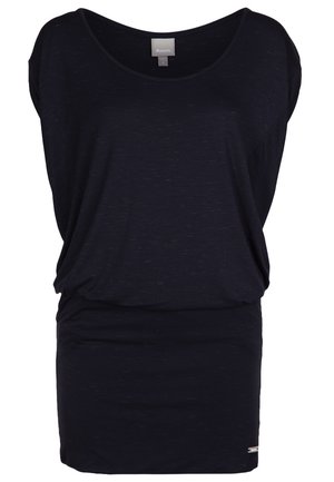 Femme portant une robe courte bleu marine, des baskets blanches et tenant un sac à dos en cuir noir à franges d'une main.