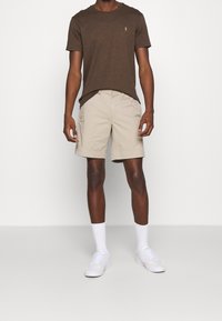 Brun kortärmad t-shirt med en liten logotyp, kombinerad med beige shorts och vita sneakers. Modellen står med armarna avslappnade längs sidorna.