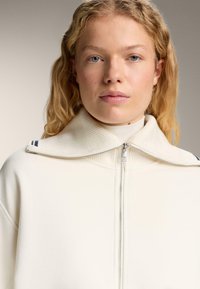 O femeie blondă cu piele deschisă poartă un pulover crem cu fermoar și guler gros cu nervuri, stând în fața unui fundal gri simplu.