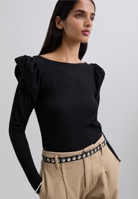 Femme portant un haut noir texturé à manches longues avec épaules bouffantes, un pantalon beige et une ceinture noire avec des rivets en argent, les mains dans les poches.