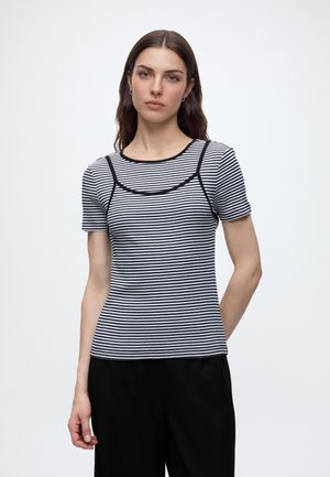 Jeune femme aux longs cheveux bruns portant un haut à manches courtes rayé noir et blanc à rayures horizontales et un pantalon noir, debout devant un fond blanc.