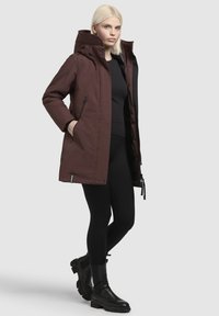 Bordeauxrode parka met een capuchon, twee zijzakken, zwarte ritsen en verstelbare trekkoorden. Gedragen over een zwart topje en gecombineerd met zwarte leggings en laarzen.