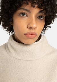 Joven con cabello rizado y oscuro, vistiendo un suéter de felpa crema con cuello alto, mirando al frente con una expresión neutral.