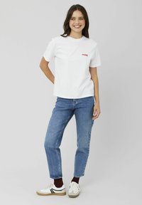 T-shirt en coton blanc avec un graphique rouge "MORE" sur la poitrine, associé à un jean bleu taille haute et des baskets blanches avec des rayures noires.