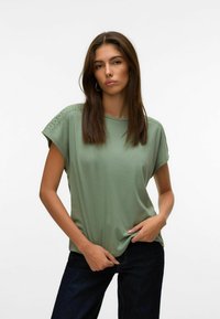 Žalia trumpomis rankovėmis palaidinė su laisvu kirpimu, su nėrinių detalėmis ant pečių. Medžiaga atrodo minkšta ir lengva. Sukomplektuota su tamsiomis džinsais.