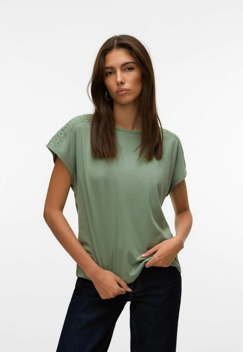 Žalia trumpomis rankovėmis palaidinė su laisvu kirpimu, su nėrinių detalėmis ant pečių. Medžiaga atrodo minkšta ir lengva. Sukomplektuota su tamsiomis džinsais.