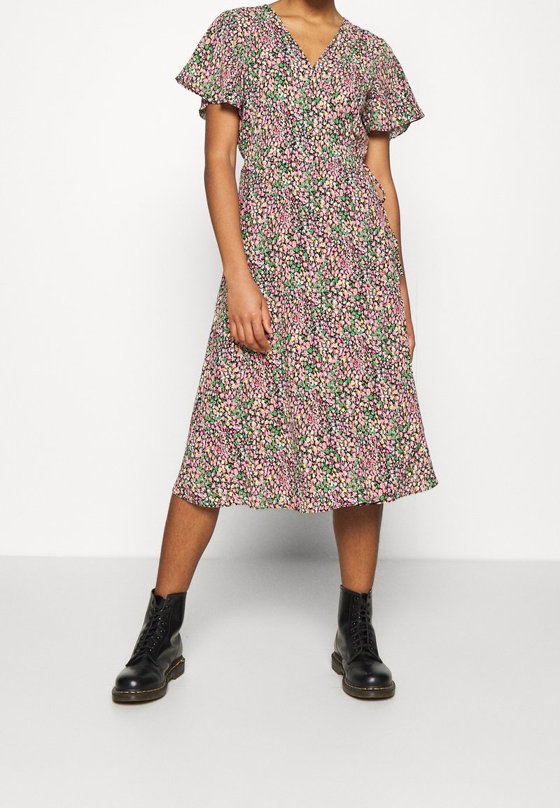 Blumenkleid aus pinkem, grünem und weißem Stoff mit V-Ausschnitt und kurzen, schwingenden Ärmeln. Knielange Länge, kombiniert mit schwarzen Schnürstiefeln.