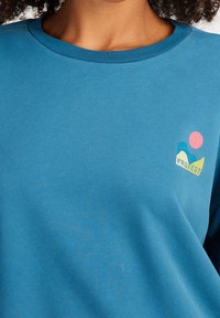 Camicia a maniche lunghe in cotone blu con collo a girocollo, caratterizzata da un logo multicolore che raffigura onde e un sole sul lato sinistro del petto.