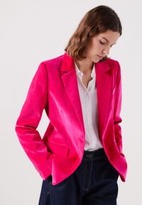 Blazer de terciopelo rosa con solapas rectas, dos bolsillos frontales y detalles de botones, vestido sobre una blusa blanca y pantalones de mezclilla oscura.