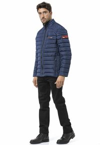 Doudoune bleu marine avec coutures verticales, col montant, deux poches latérales zippées et patchs de marque. Portée avec un jean noir et des bottes.