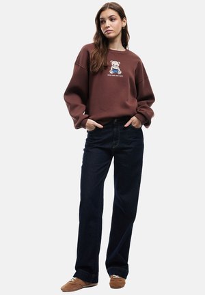 Brun sweatshirt med et bjørnemotiv og teksten "hold dig cool og rolig". Båret med mørkeblå jeans og brune sko, med en afslappet pasform.