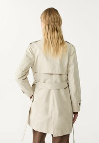Beżowy płaszcz trench z paskiem w talii, pagonami na ramionach i długimi rękawami. Gładka tekstura, projekt do kolan, minimalistyczny styl.