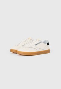 White sneakers with textured upper, gray accents, and a gum sole. Features flat laces and a logo near the tongue. Black heel tab detail.