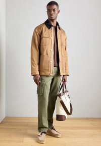 Brun bomuldsjakke med sort krave, to brystlommer, olivengrønne cargo bukser, beige mokkasiner og en beige tote taske med brune accenter.
