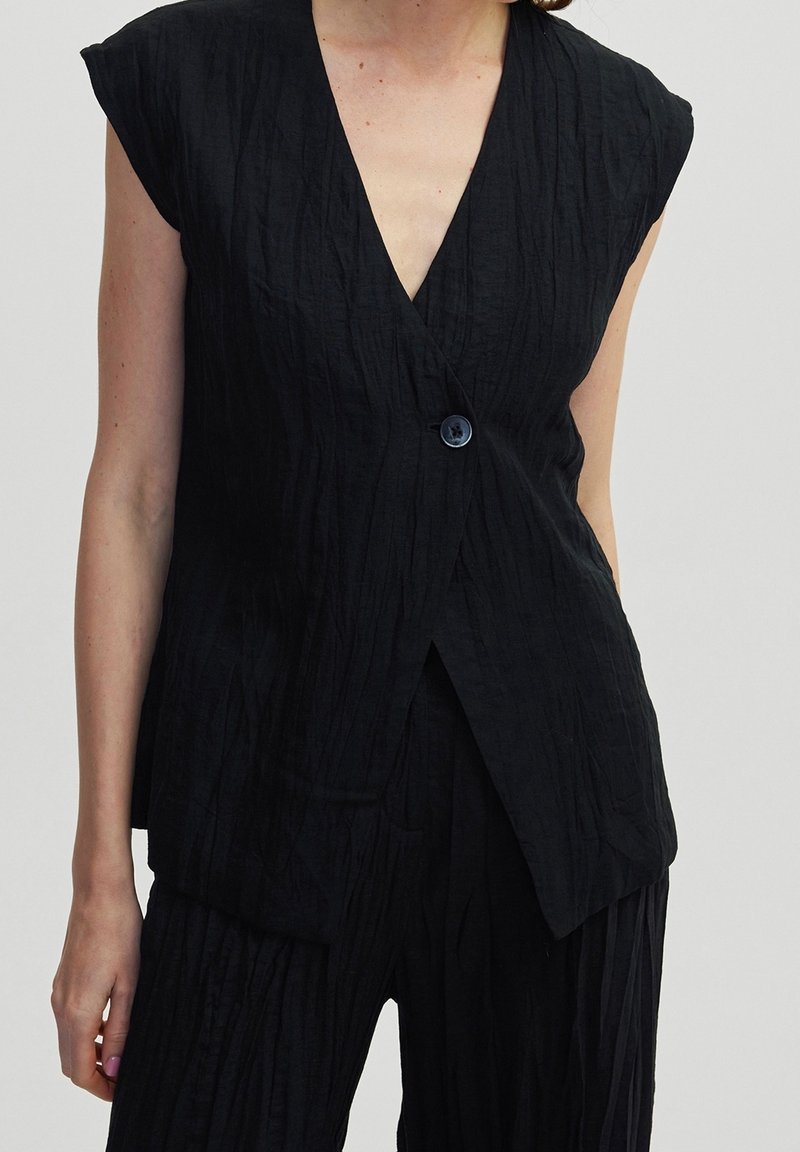 Femme portant un haut cache-cœur noir texturé sans manches avec un seul bouton et un pantalon noir texturé assorti.