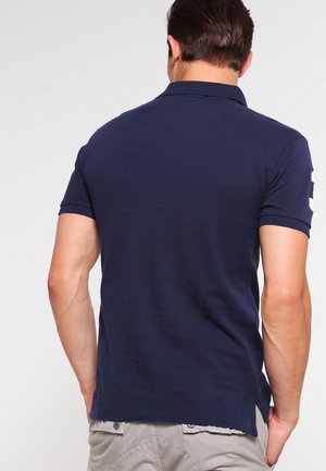 Homme portant un polo manches courtes bleu marine et un pantalon gris clair, vu de dos sur un fond uni.