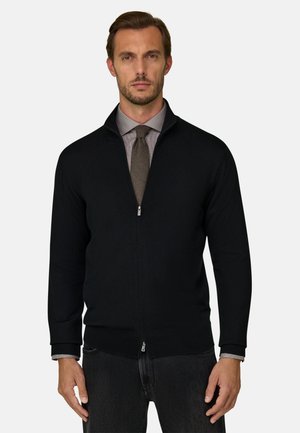 Homme portant un pull zippé noir sur une chemise grise et une cravate marron, debout devant un fond clair uni.