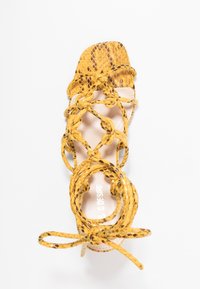 Żółte sandały w wężowy wzór z płaską podeszwą, z plecionymi paskami owijającymi stopę oraz kwadratowym noskiem.