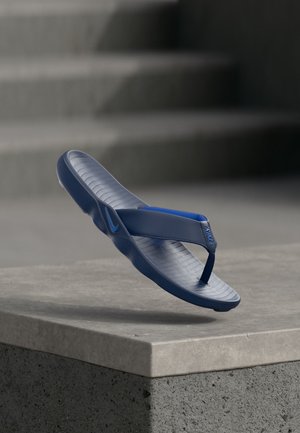 Μπλε σαγιονάρα Nike που αιωρείται πάνω από μια τσιμεντένια γωνία με υφή στο πέλμα και λουράκι με το λογότυπο σε απαλό φωτισμό.