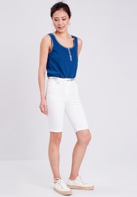 Top senza maniche blu con bottoni frontali, abbinato a pantaloni corti bianchi fino al ginocchio. Il modello indossa scarpe da ginnastica bianche con dettagli texture.