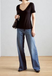 Frau trägt ein schwarzes, gewelltes V-Ausschnitt-Oberteil, weit geschnittene blaue Jeans, schwarze Schuhe, hält eine schwarze Schultertasche und steht auf einem Holzboden vor einer grauen Wand.