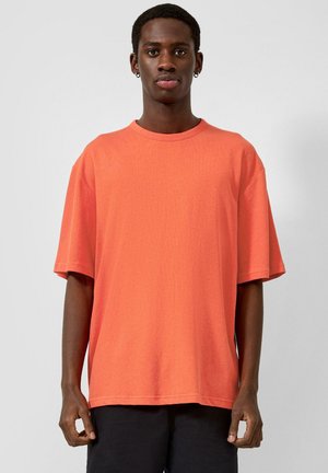 Jeune homme portant un t-shirt orange surdimensionné à manches courtes et un pantalon noir, debout devant un fond gris clair uni.
