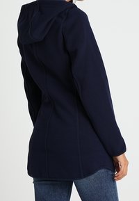 Laivastonsininen hupullinen takki, jossa on teksturoitu kangas, istuva malli ja kontrastisaumaukset. Siinä on pitkä leikkaus ja sivutaskut.