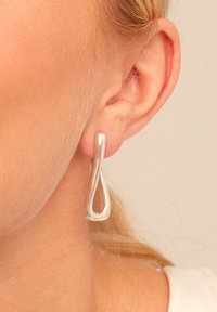 Pendientes colgantes de plata con un diseño escultórico en espiral, que presentan un acabado liso y pulido y una forma de lágrima, colocados junto a la oreja.