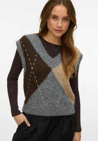 Chaleco de punto en tonos gris, marrón y beige con un patrón de diamantes, textura esponjosa, sisas anchas y detalles de puntadas visibles, combinado sobre una camiseta oscura.