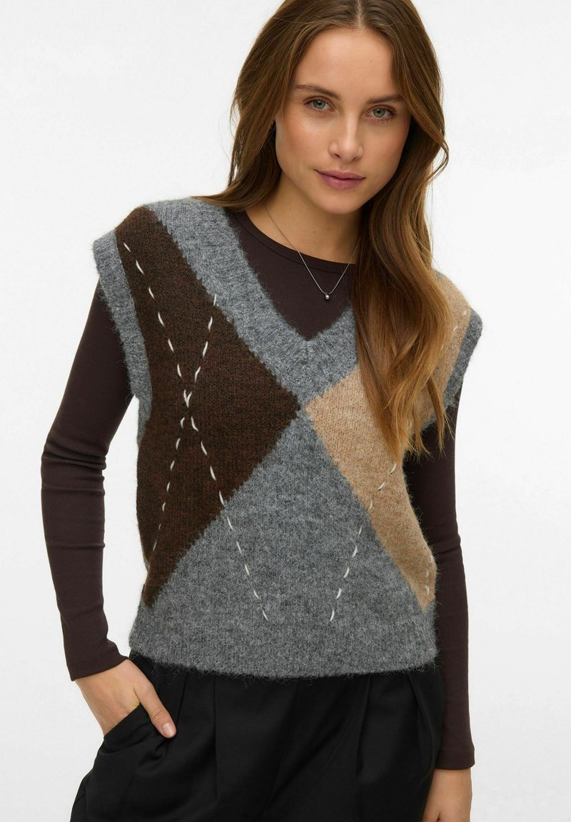 Chaleco de punto en tonos gris, marrón y beige con un patrón de diamantes, textura esponjosa, sisas anchas y detalles de puntadas visibles, combinado sobre una camiseta oscura.