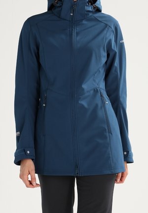 Kobieta ubrana w niebieską kurtkę z kapturem zapinaną na zamek błyskawiczny, z zamykanymi na zamek kieszeniami po bokach i regulowanymi mankietami, stojąca na jednolitym tle.