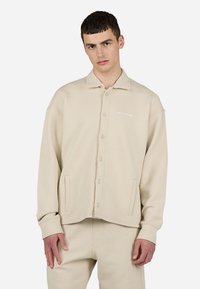Beige button-up jacket in textured fabric, featuring a pointed collar, two side pockets, and a logo on the chest.
