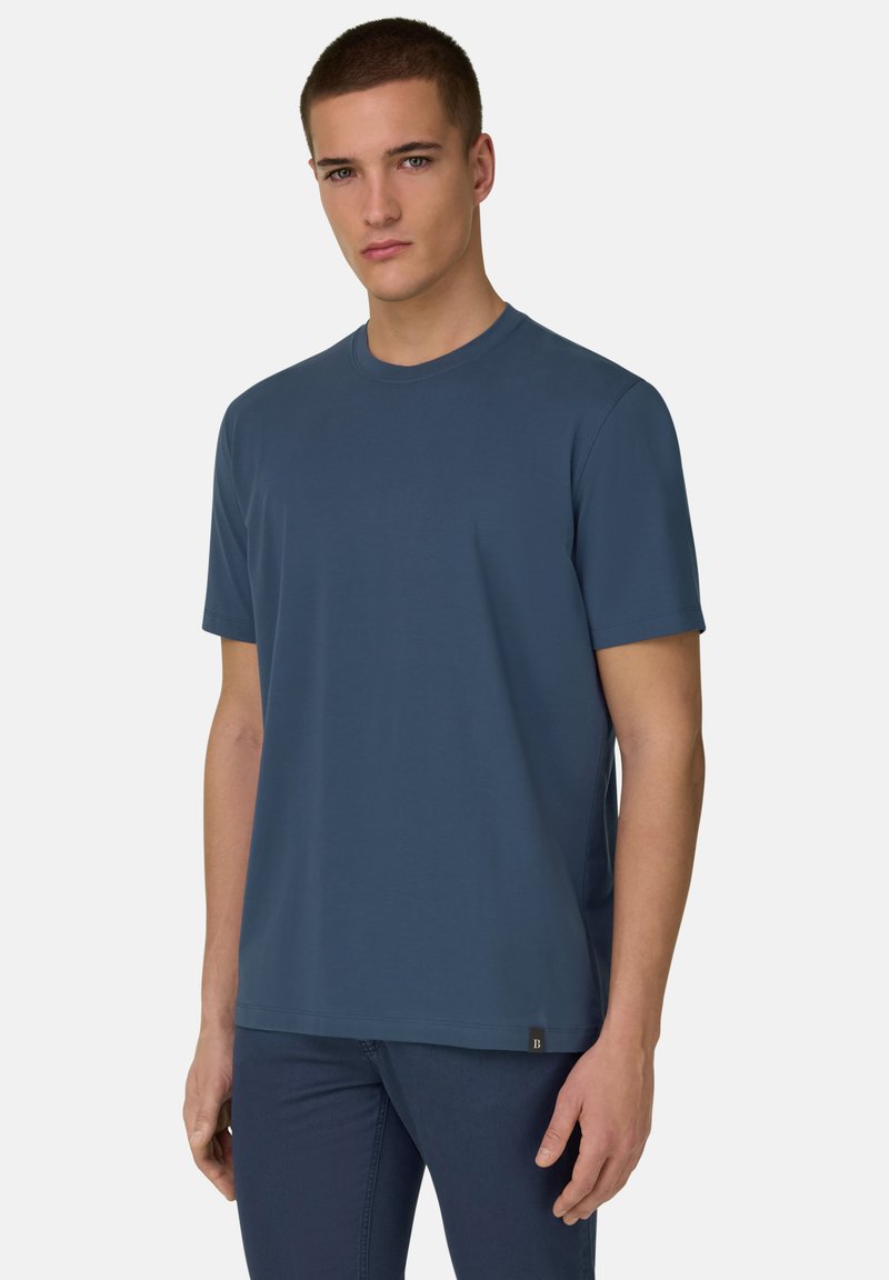 Jeune homme portant un t-shirt bleu uni à manches courtes et un pantalon sombre, debout devant un fond gris clair.