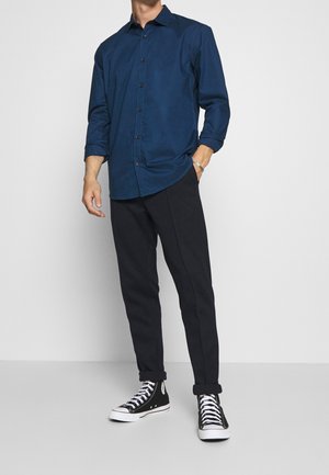 Laivastonsininen napillinen paita, jossa pitkät hihat, yhdistetty mustiin kapeneviin housuihin ja mustiin korkeavartisiin tennareihin. Paidassa on klassinen kaulus.