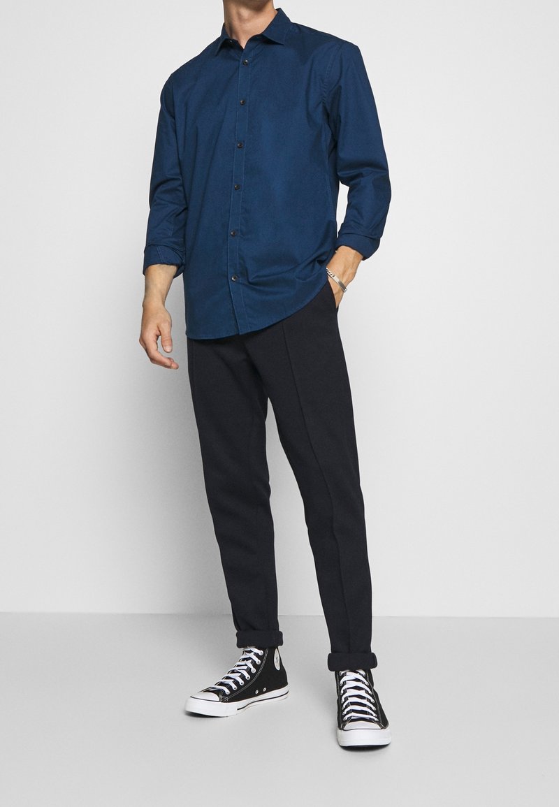 Camisa azul marino abotonada con mangas largas, combinada con pantalones negros entallados y zapatillas negras de caña alta. La camisa tiene un cuello clásico.