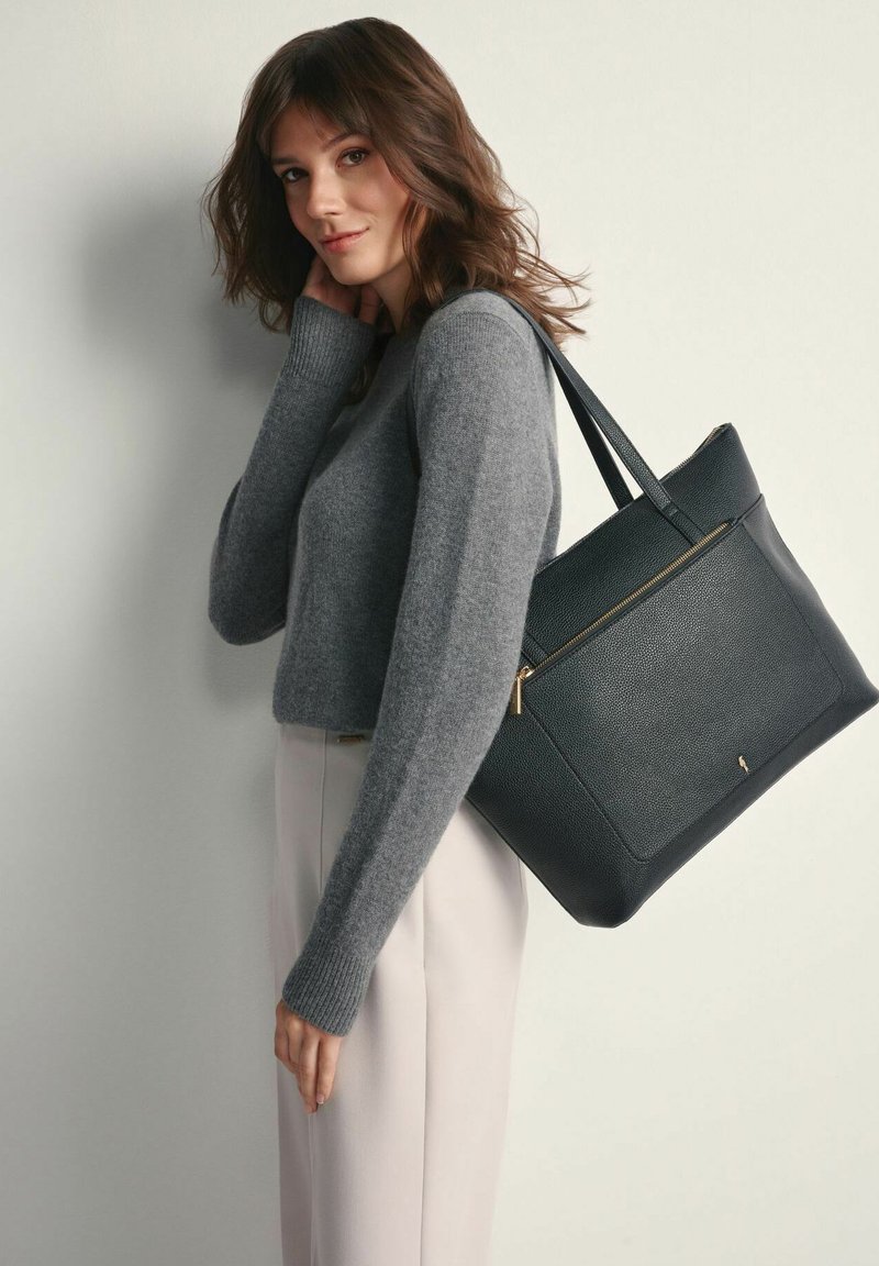 Czarna torba tote z teksturowanej skórki, z złotym zamkiem błyskawicznym, połączona z szarym swetrem dzianinowym i jasnymi spodniami, trzymana przez osobę na ramieniu.