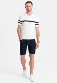 Camisa polo blanca con rayas marinas, combinada con pantalones cortos de mezclilla oscuros y zapatillas blancas. El modelo está de pie con una mano en el bolsillo, con un fondo neutro.