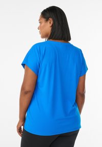 Frau mit schulterlangem schwarzen Haar, die ein leuchtend blaues Kurzarmshirt und schwarze Hosen trägt, von hinten vor einem weißen Hintergrund.