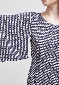 Námořnické a bílé šaty s potiskem, které mají krátké, široké rukávy. Tkanina se vyznačuje geometrickým vzorem s malými tvary a jemnou texturou.