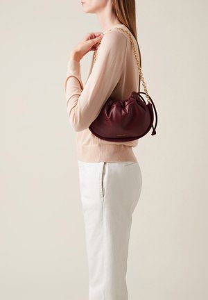 Burgundy leather handbag with gathered top, gold chain strap, and woven accents. Styled with a light sweater and pale trousers.